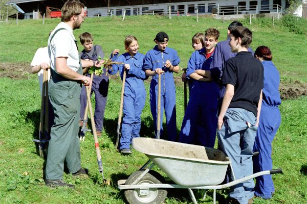 Teachers, students of a vocational and educational training school standing on a meadow, in the foreground a wheelbarrow