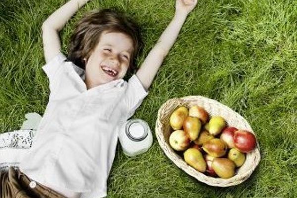 A laughing girl in a white blouse is lying on the green meadow with a bottle of milk and a basket of apples next to her