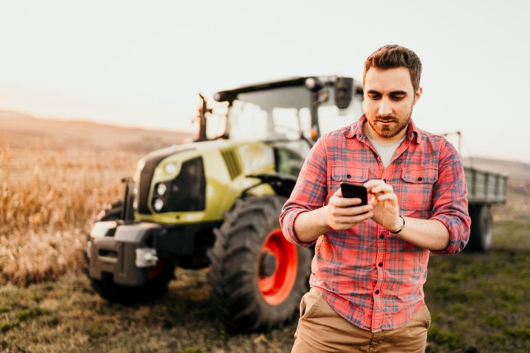 Ein Landwirt am Feld mit Handy, Hintergrund Traktor