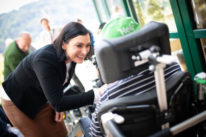 Bundesministerin Köstinger lächelt und spricht mit einer Person in einem Rollstuhl; sichtbar ist nur die Rückseite des Rollstuhls